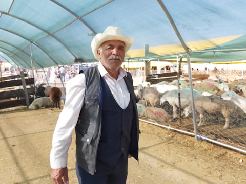 An older man with a white mustache and beard is wearing a cowboy hat, a white shirt, and a dark vest. He is standing inside a covered area that appears to be a livestock market. Behind him, sheep are enclosed within a fenced pen, and the ground is covered with straw and dirt. The roof is made of semi-transparent blue and yellow material, letting in some sunlight.