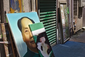 An alleyway with several framed paintings leaning against a wall. One painting prominently features the face of a man with dark hair and another painting partially visible features a panda. Green shutters partially conceal another piece of artwork, and a textured wall runs alongside.