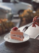 A slice of sponge cake with a light pink color, filled with cream and strawberries, sits on a decorative white plate. A hand is holding a fork, poised to take a bite. The background appears blurred, indicating an outdoor setting, possibly a café or patio.