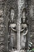 Intricately carved stone relief depicting two standing figures with ornate headdresses and traditional attire. The figures are surrounded by elaborate, decorative patterns etched into the stone background, showcasing ancient artistry.