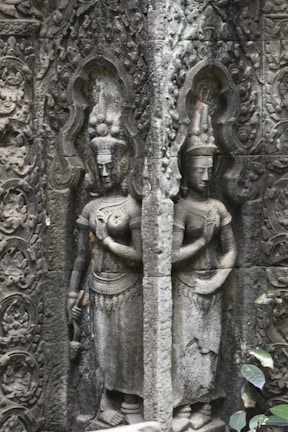 Intricately carved stone relief depicting two standing figures with ornate headdresses and traditional attire. The figures are surrounded by elaborate, decorative patterns etched into the stone background, showcasing ancient artistry.