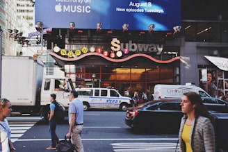 A busy urban street scene featuring a subway entrance with signage displaying various train lines. Several vehicles, including a truck and a police van, are on the street, while pedestrians walk on the sidewalk. Above the subway entrance is a large billboard advertising Apple Music.