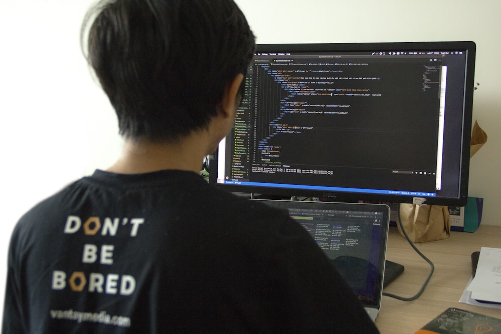 man in black shirt using laptop computer and flat screen monitor