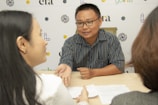 Three people are engaged in a conversation at a table. The man, wearing a striped shirt and glasses, is gesturing with his hand while speaking to the two women seated across from him. Documents are placed on the table. A branded backdrop is visible in the background.