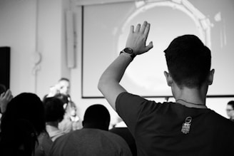 A group of people are gathered in a room facing a speaker at the front. One person in the foreground raises their hand, while the others seem to be listening intently. The atmosphere is akin to a classroom or seminar setting. The scene is captured in black and white, adding a timeless and serious tone to the moment.