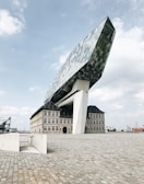 A modern architectural structure featuring a large, angular glass and steel form perched atop a historic brick building. The sky is partly cloudy, and the ground is paved with cobblestones.