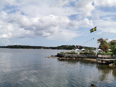 A scenic view of a calm body of water with a rocky shoreline. There is a small island or peninsula with greenery and trees. A Swedish flag is prominently displayed on a flagpole, along with a string of small colorful nautical flags. A few open-air umbrellas are visible, suggesting a seating area or café.