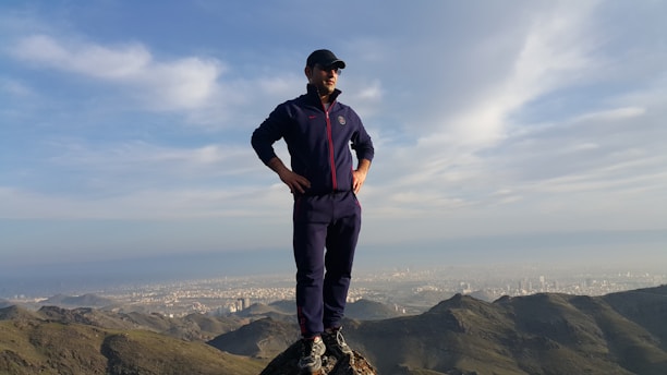 A person stands confidently on a mountain peak overlooking a sprawling urban landscape in the distance. The individual is wearing athletic clothing and a cap, framed by a vast blue sky with scattered clouds.