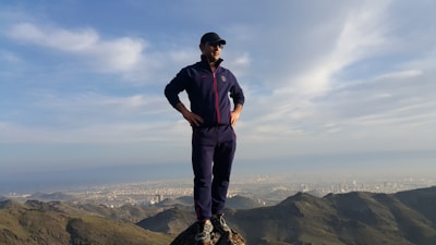 A person stands confidently on a mountain peak overlooking a sprawling urban landscape in the distance. The individual is wearing athletic clothing and a cap, framed by a vast blue sky with scattered clouds.