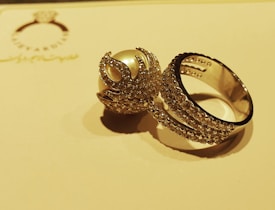 A close-up of a silver ring adorned with numerous sparkling gemstones is displayed on a light, golden-colored surface. The ring features an intricate design, including a large, pearl-like gemstone encased in decorative metalwork.