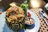 A dish of pineapple fried rice is served in a pineapple shell, garnished with onions and herbs. There is a small bowl of sauce with sliced chili and a side of fresh vegetables on the same plate. The setup includes a placemat and a spoon and fork beside the plate.