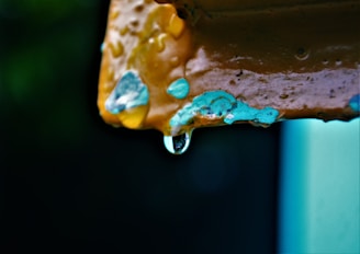 A close-up of a rusted metal surface with peeling paint, featuring a droplet of water hanging from the edge. The chipped paint reveals layers of different colors, including blue and yellow, highlighted against a dark background.