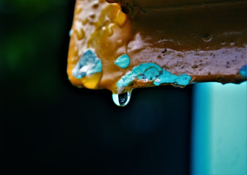 A close-up of a rusted metal surface with peeling paint, featuring a droplet of water hanging from the edge. The chipped paint reveals layers of different colors, including blue and yellow, highlighted against a dark background.