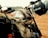 A close-up view of a vintage motorcycle parked on a dirt track. A khaki canvas bag is attached to the motorcycle, holding a bottled drink. A black helmet with the words 'Do Fun' is hanging on the handlebar. The background is slightly blurred, indicating other motorcycles and a vast open space.