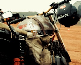 A close-up view of a vintage motorcycle parked on a dirt track. A khaki canvas bag is attached to the motorcycle, holding a bottled drink. A black helmet with the words 'Do Fun' is hanging on the handlebar. The background is slightly blurred, indicating other motorcycles and a vast open space.