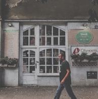 A man is walking past an old building with a large white, vintage-style door and multiple small window panes. The facade has signs indicating a cafe named 'Fresh Food' and a hostel called 'City Hostel'. Potted plants adorn the front, and the building shows signs of wear, such as peeling paint.