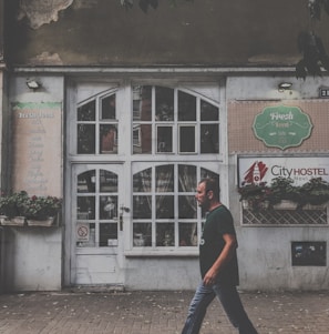 A man is walking past an old building with a large white, vintage-style door and multiple small window panes. The facade has signs indicating a cafe named 'Fresh Food' and a hostel called 'City Hostel'. Potted plants adorn the front, and the building shows signs of wear, such as peeling paint.