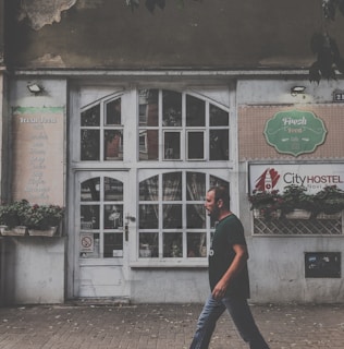 A man is walking past an old building with a large white, vintage-style door and multiple small window panes. The facade has signs indicating a cafe named 'Fresh Food' and a hostel called 'City Hostel'. Potted plants adorn the front, and the building shows signs of wear, such as peeling paint.