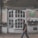 A man is walking past an old building with a large white, vintage-style door and multiple small window panes. The facade has signs indicating a cafe named 'Fresh Food' and a hostel called 'City Hostel'. Potted plants adorn the front, and the building shows signs of wear, such as peeling paint.