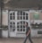A man is walking past an old building with a large white, vintage-style door and multiple small window panes. The facade has signs indicating a cafe named 'Fresh Food' and a hostel called 'City Hostel'. Potted plants adorn the front, and the building shows signs of wear, such as peeling paint.