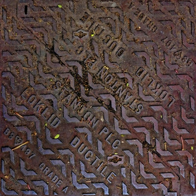 A metal manhole cover with textured patterns and embossed text, partially covered in dirt and small leaves. The text includes the words 'Stanton PLC' and 'Ductile' along with other manufacturing details.