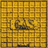 A yellow metal panel with a grid pattern and raised text that reads 'GAS' in the center, surrounded by signs of weathering and dirt.