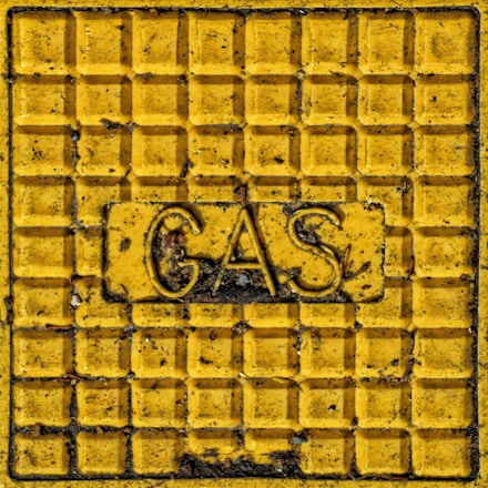 A yellow metal panel with a grid pattern and raised text that reads 'GAS' in the center, surrounded by signs of weathering and dirt.