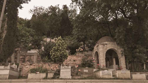 A historic cemetery surrounded by lush greenery, featuring old gravestones and an ornate stone structure with a domed roof. The area is densely planted with trees and bushes, adding a serene and solemn atmosphere to the landscape.