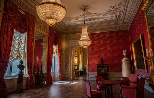 An opulently decorated room featuring red and gold wallpaper with intricate patterns. A large chandelier hangs from a lavishly detailed ceiling. The room is furnished with elegant red upholstered chairs and a circular table. Tall draped curtains frame the windows, and several decorative busts and candelabras adorn the space. The atmosphere is one of classical luxury.