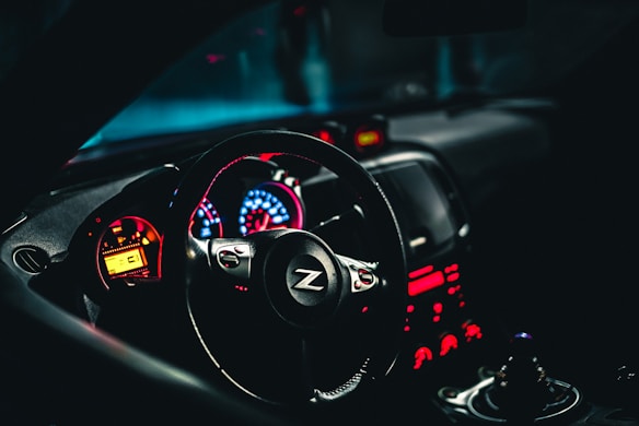 A car interior highlighting a sleek steering wheel with a 'Z' logo and an illuminated dashboard displaying vibrant red and blue lights. The design gives a modern and dynamic feel, with the gearshift visible in the foreground.