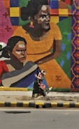 A person in traditional dance attire is performing an expressive dance move in front of a vibrant mural on a wall. The mural features large, colorful geometric shapes and stylized portraits of people in warm tones. The scene is set against an urban background with a road and sidewalk in the foreground.