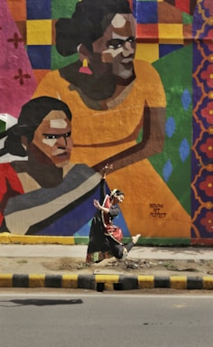 A person in traditional dance attire is performing an expressive dance move in front of a vibrant mural on a wall. The mural features large, colorful geometric shapes and stylized portraits of people in warm tones. The scene is set against an urban background with a road and sidewalk in the foreground.