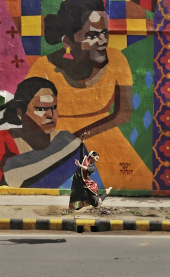 A person in traditional dance attire is performing an expressive dance move in front of a vibrant mural on a wall. The mural features large, colorful geometric shapes and stylized portraits of people in warm tones. The scene is set against an urban background with a road and sidewalk in the foreground.