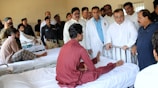 A group of medical professionals and possibly officials are gathered around a patient in a hospital ward. The patient is seated on a hospital bed, engaging with the people around him. Several men in uniform and plain clothes are present, suggesting a formal visit or inspection.