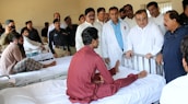 A group of medical professionals and possibly officials are gathered around a patient in a hospital ward. The patient is seated on a hospital bed, engaging with the people around him. Several men in uniform and plain clothes are present, suggesting a formal visit or inspection.