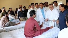 A group of medical professionals and possibly officials are gathered around a patient in a hospital ward. The patient is seated on a hospital bed, engaging with the people around him. Several men in uniform and plain clothes are present, suggesting a formal visit or inspection.