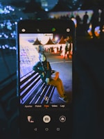 A smartphone captures an image of a person sitting on a bench in a brightly lit outdoor setting with decorative lights. The background features several triangular tents and people walking around, creating a festive atmosphere.