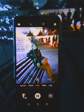A smartphone captures an image of a person sitting on a bench in a brightly lit outdoor setting with decorative lights. The background features several triangular tents and people walking around, creating a festive atmosphere.