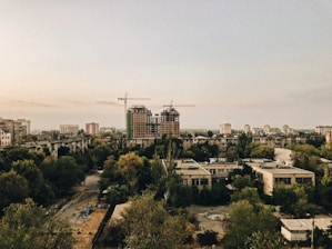 An urban scene with several partially constructed high-rise buildings, surrounded by greenery and smaller residential structures. Two cranes are visible on top of one of the buildings, indicating ongoing construction. The sky is overcast, creating a muted and subdued lighting over the cityscape. Trees with green and yellow foliage dominate the foreground, suggesting a city in transition between seasons.