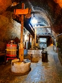 A dimly lit underground cellar featuring a large wooden wine press. The stone flooring and arched brick ceiling add a rustic feel to the setting. Various winemaking equipment, including a metal barrel and a red basket press, are visible.
