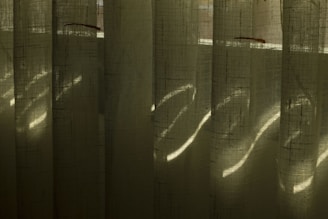 Vertical blinds made of fabric with light filtering through, creating patterns of light and shadow. The fabric has a textured, translucent quality, and the light appears to create subtle striations across the surface.