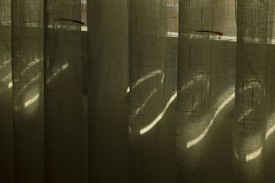 Vertical blinds made of fabric with light filtering through, creating patterns of light and shadow. The fabric has a textured, translucent quality, and the light appears to create subtle striations across the surface.