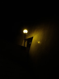 A bright street lamp illuminates a dark area, casting a warm yellow glow. The light appears against a tiled wall, creating a textured background. The scene is primarily dark, with the lamp providing the main source of light.