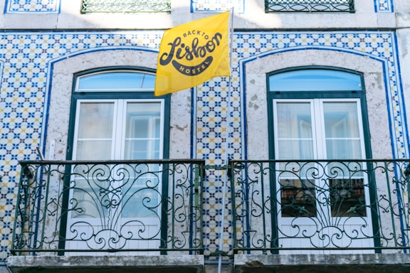 A building facade featuring intricate blue and white tile patterns typical of Portuguese architecture, with two windows framed by ornate wrought iron railings. A bright yellow flag with the text 'Back to Lisbon Hostel' adds a vibrant touch to the scene.