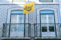 A building facade featuring intricate blue and white tile patterns typical of Portuguese architecture, with two windows framed by ornate wrought iron railings. A bright yellow flag with the text 'Back to Lisbon Hostel' adds a vibrant touch to the scene.