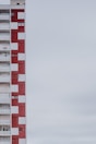 A multi-story apartment building with a distinctive red and white checkered facade. The structure is positioned on the left side of the image, leaving a large expanse of overcast sky to the right. The building features balconies, windows, and a sign indicating something is for rent or sale.