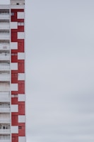 A multi-story apartment building with a distinctive red and white checkered facade. The structure is positioned on the left side of the image, leaving a large expanse of overcast sky to the right. The building features balconies, windows, and a sign indicating something is for rent or sale.