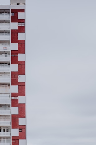 A multi-story apartment building with a distinctive red and white checkered facade. The structure is positioned on the left side of the image, leaving a large expanse of overcast sky to the right. The building features balconies, windows, and a sign indicating something is for rent or sale.