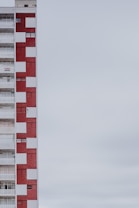 A multi-story apartment building with a distinctive red and white checkered facade. The structure is positioned on the left side of the image, leaving a large expanse of overcast sky to the right. The building features balconies, windows, and a sign indicating something is for rent or sale.