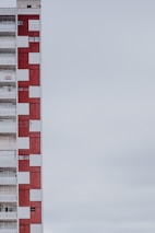 A multi-story apartment building with a distinctive red and white checkered facade. The structure is positioned on the left side of the image, leaving a large expanse of overcast sky to the right. The building features balconies, windows, and a sign indicating something is for rent or sale.